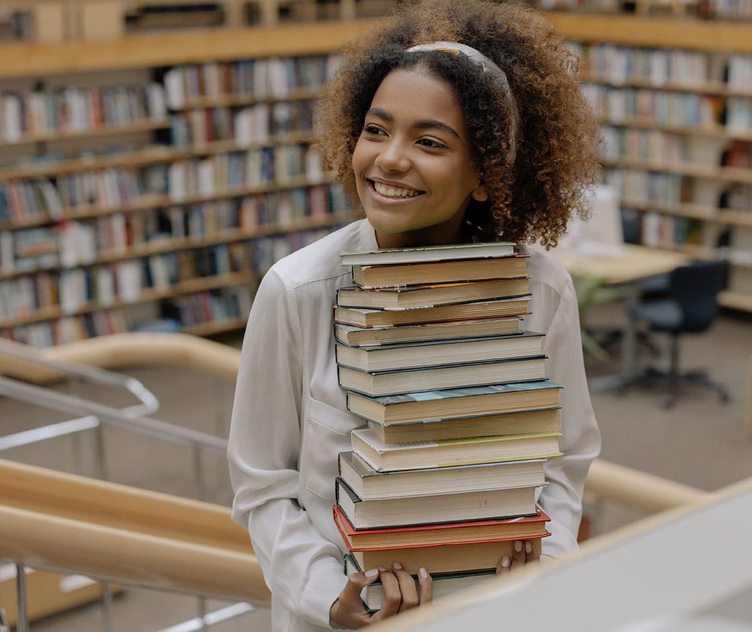 Young girl in library holding large stack of books